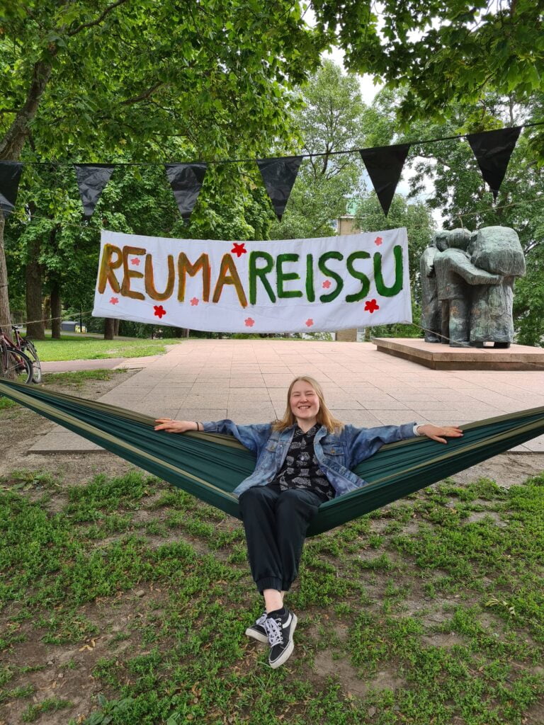 Nuori nainen istuu riippumatossa yläpuolellaan banderolli jossa teksti reumareissu.
