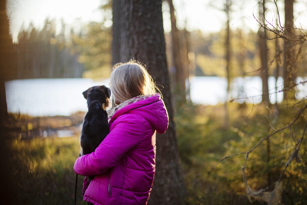 Pinkkiin takkiin pukeutunut nainen seisoo järven reunalla ja pitää sylissään pientä koiraa.