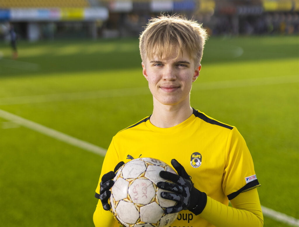 Nuori mies keltaisessa pelipaidassa pitää jalkapalloa kädessään.