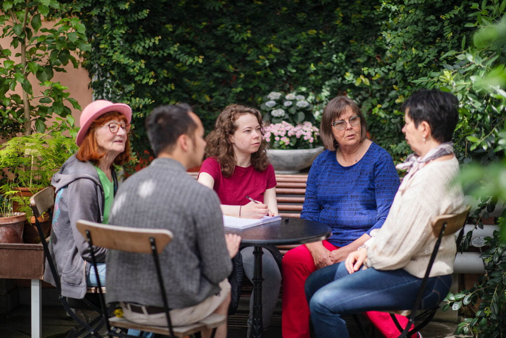Vertaisryhmä istuu vehreässä puutarhassa pöytäryhmän ympärillä ja kuuntelee tarkasti kun yksi paikalla olevista puhuu.