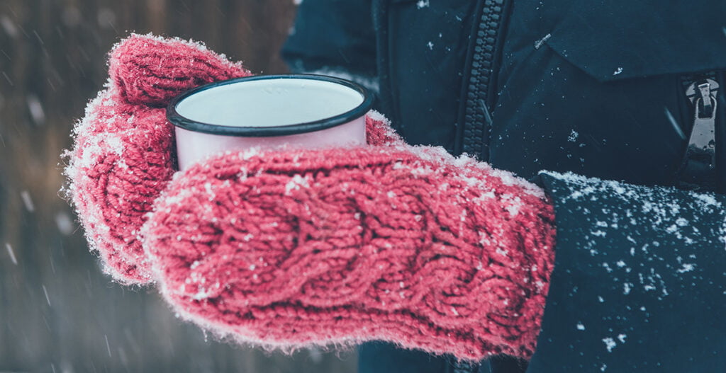 henkilö pitelee lumisateessa lapaset kädessään kahvikuppia.