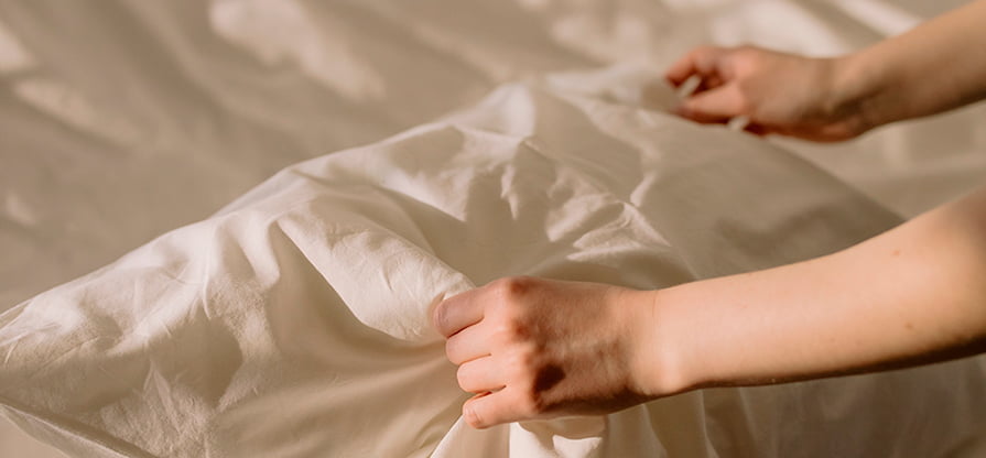 Kuvassa näkyy valkoiset lakanat ja tyyny ja kädet petaamassa sänkyä
