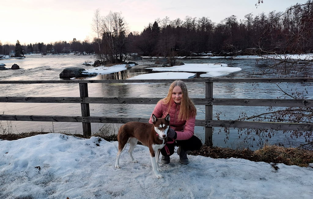 Nuori nainen poseeraa koiran kanssa kameralle talvisen joen reunalla.