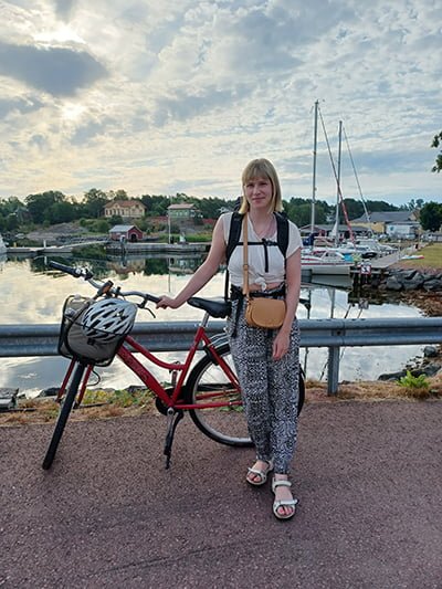Nuori nainen seisoo pyörän kanssa reppu selässä, taustalla saaristomaisema.