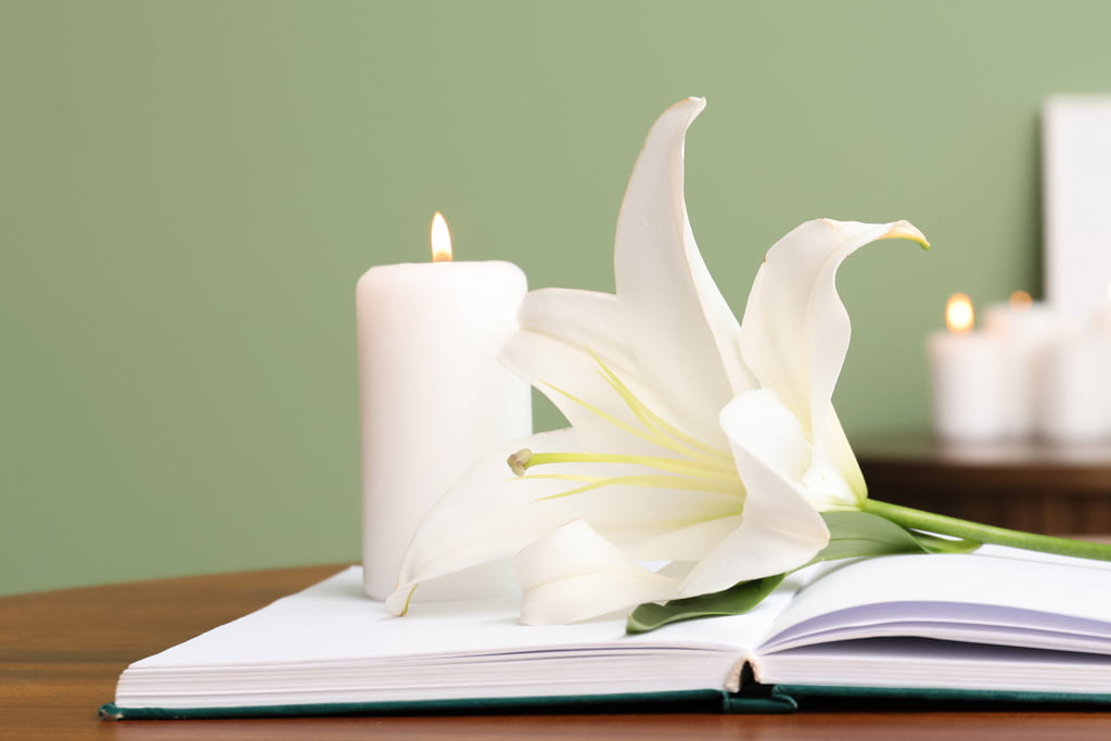 Valkoinen kynttilä, valkoinen lilja ja avoin muistokirja pöydällä.