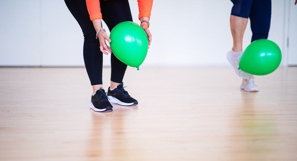 Kaksi ihmistä kurottelee ilmapalloja lattialta liikuntasalissa.