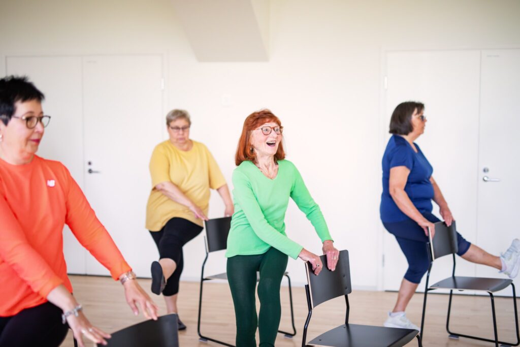 Neljä henkilöä seisoo tuolin takana ja heiluttaa toista jalkaansa hymyillen ja pitäen kiinni tuolin selkänojasta.