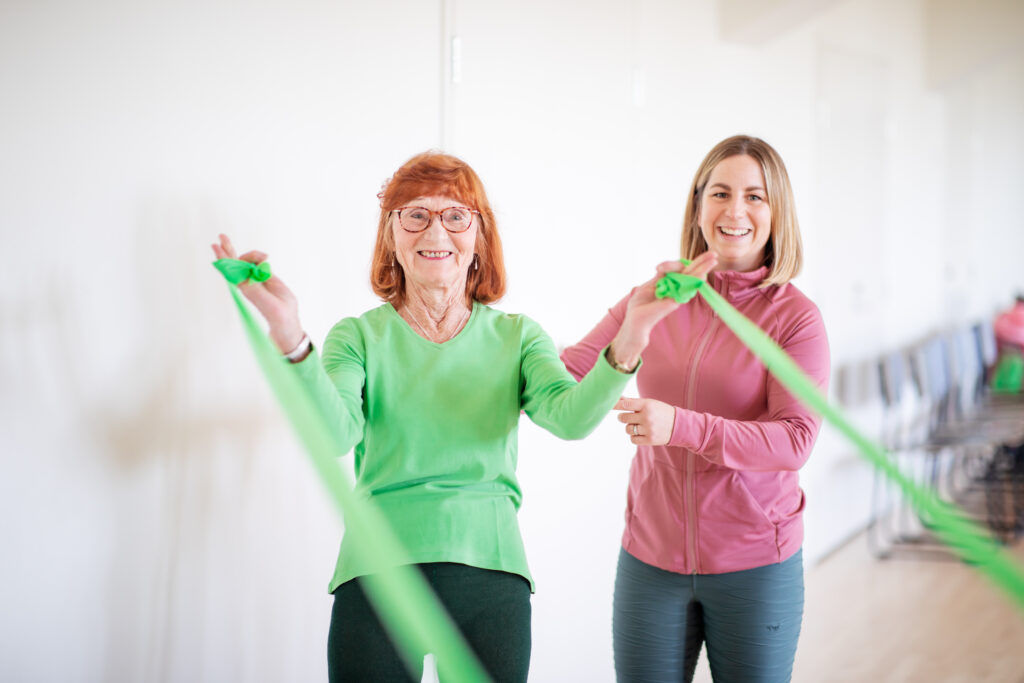 Kuvassa kaksi naisoletettua hymyilee ja, joista toinen liikkumassa vastusnauhoilla ja toinen ohjaamassa.