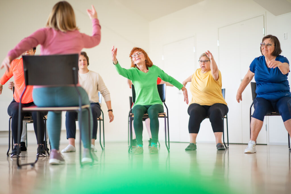 Kuvassa henkilöitä tuolijumppaamassa ohjaajan johdolla.