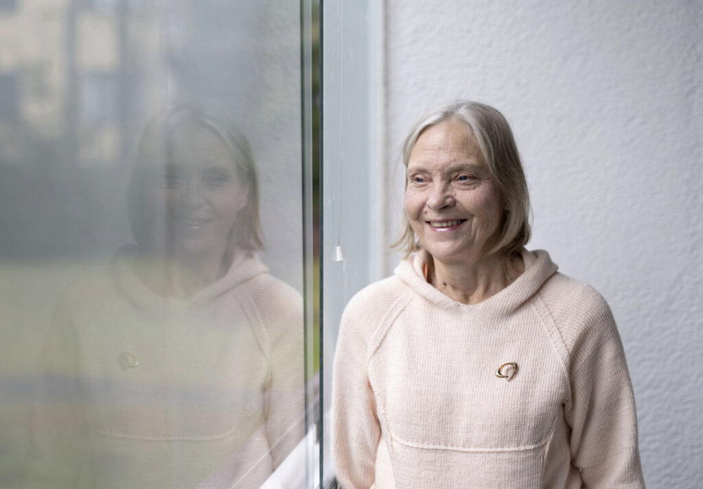 Nainen katsoo ulos ikkunasta ja hänen ikkunalasista heijastuvat kasvonsa katsovat suoraan katsojaan.