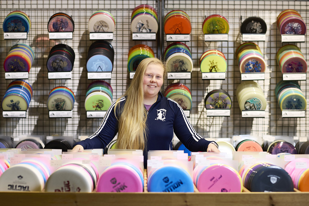 Nainen seisoo myymälässä, takana olevalla seinällä ja edessä olevalla pöydälä eri värisiä frisbeegolfkiekkoja.