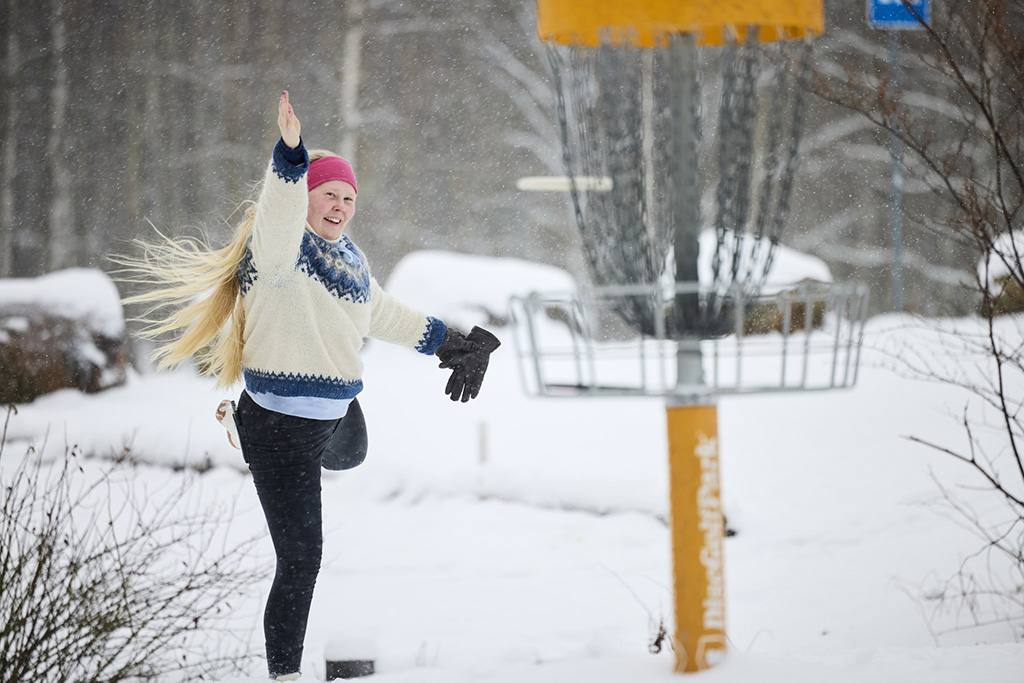 Nainen lumisessa maisemassa heittämässä frisbeetä etualalla näkyvään frisbeegolfkoriin.