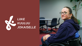 Nainen istuu pyörätuolissa toimistossa ja on kääntynyt katsomaan kameraan.